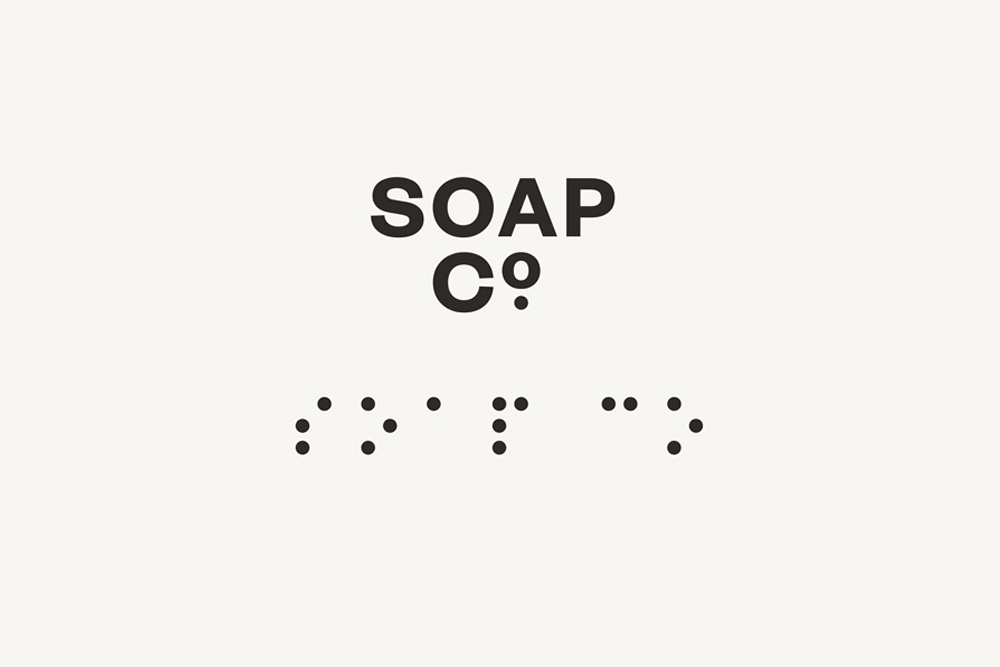 Minimalist graphic design brand identity for The Soap Co featuring centered dark typography and braille text on a pale background.