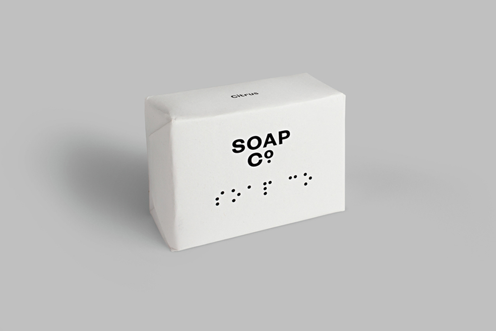 White soap bar package with black typography and braille dots on a grey seamless background