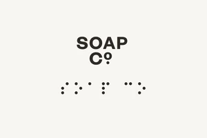 Minimalist graphic design brand identity for The Soap Co featuring centered dark typography and braille text on a pale background.
