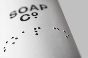 Close-up of The Soap Co. packaging showing black typography and debossed tactile braille dots on textured paper.