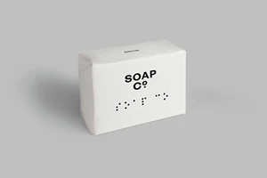 White soap bar package with black typography and braille dots on a grey seamless background