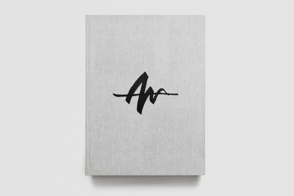 Close-up of a light grey fabric-bound book with a bold black custom logo on the cover, viewed from above against a white background.
