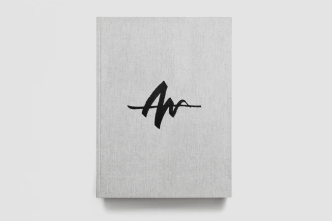 Close-up of a light grey fabric-bound book with a bold black custom logo on the cover, viewed from above against a white background.