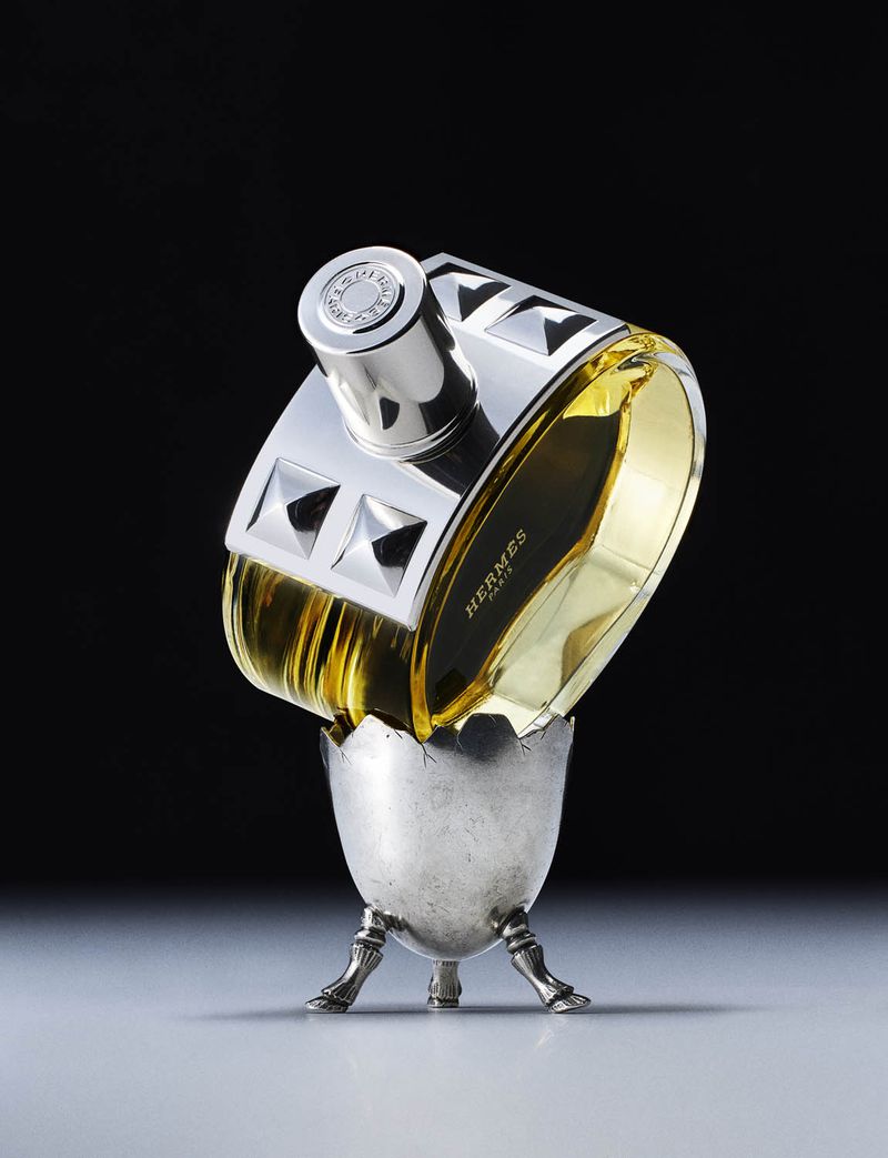 Still life studio shot of a Hermès perfume bottle with a silver decorative cap in a silver egg cup against a dark gradient background.