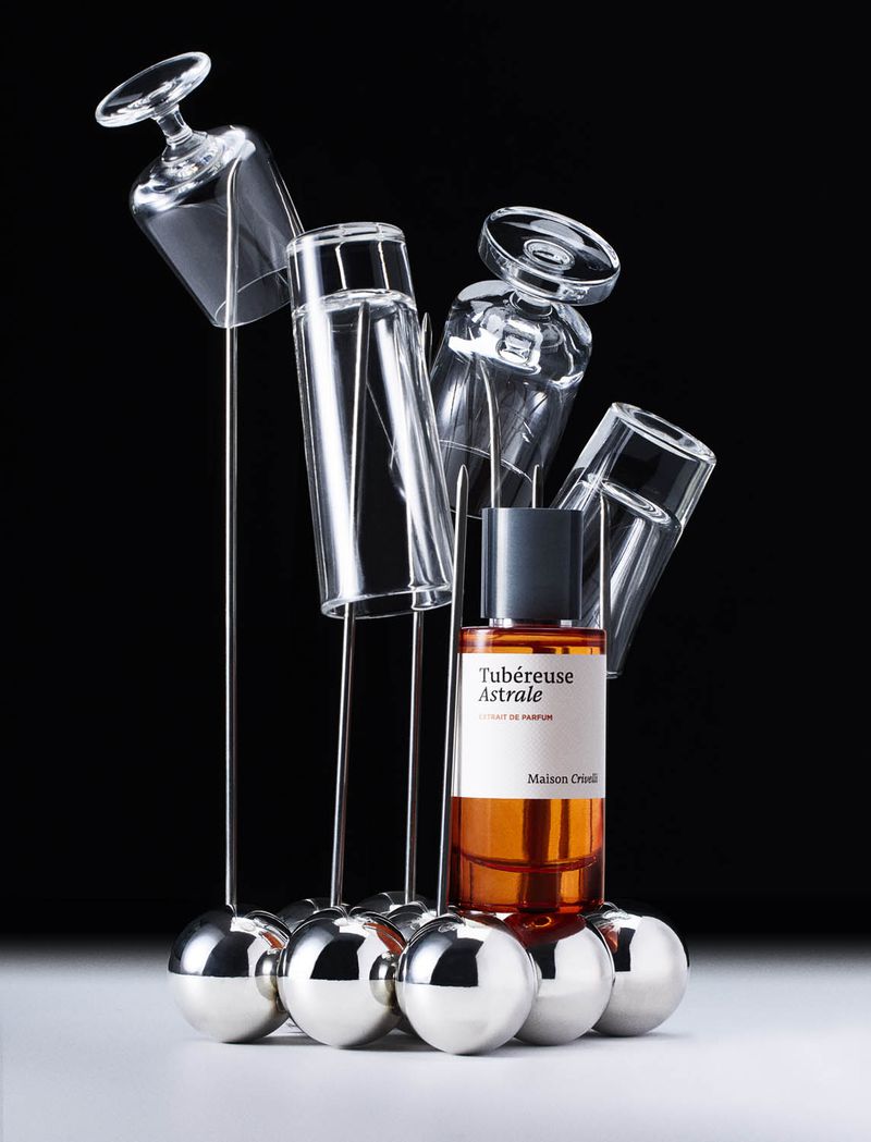 Still life shot of Maison Orieli Tubereuse Astrale perfume bottle on silver balls, surrounded by clear glassware and a black background.