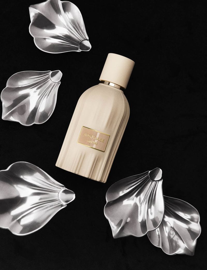 Still life studio shot of a light beige perfume bottle with gold accents surrounded by metallic silver shell-shaped objects on black backdrop.