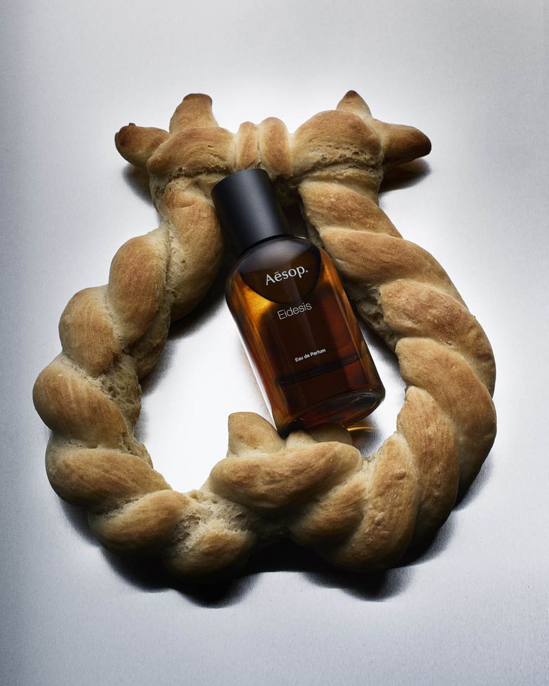 Studio shot of Aesop Eidesis perfume bottle nestled in a braided bread ring on a shiny, neutral surface with bright, even lighting.