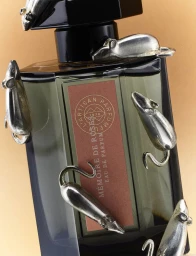 Close-up of a dark glass perfume bottle adorned with small silver metallic mouse sculptures climbing its surface against a warm tan background.
