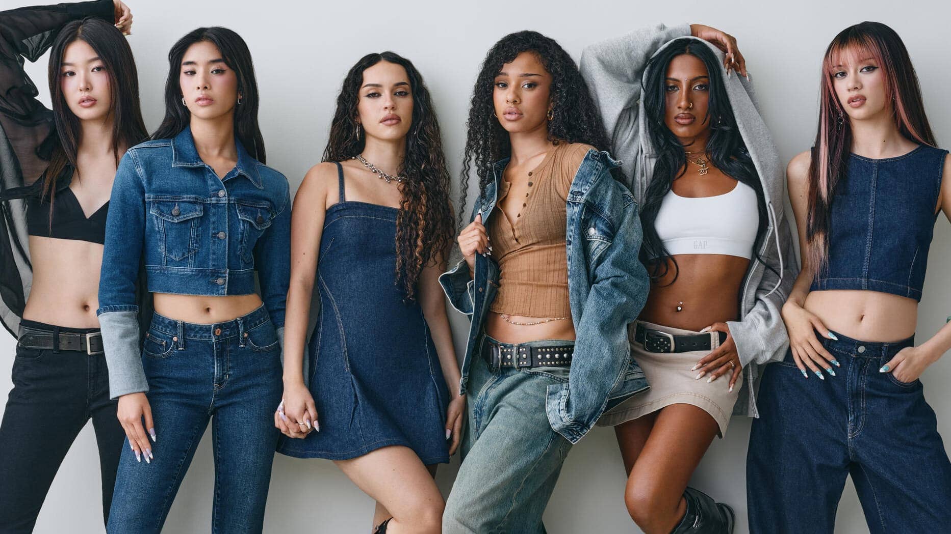 Group of six young women posing in casual denim and streetwear against a neutral light grey studio backdrop.