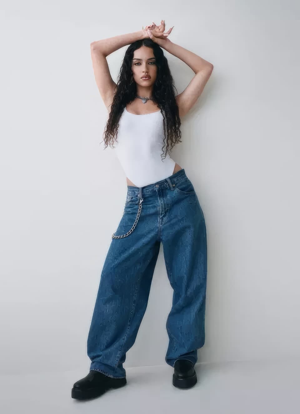 Model wearing a white tank top and blue denim jeans standing against a plain white studio background.