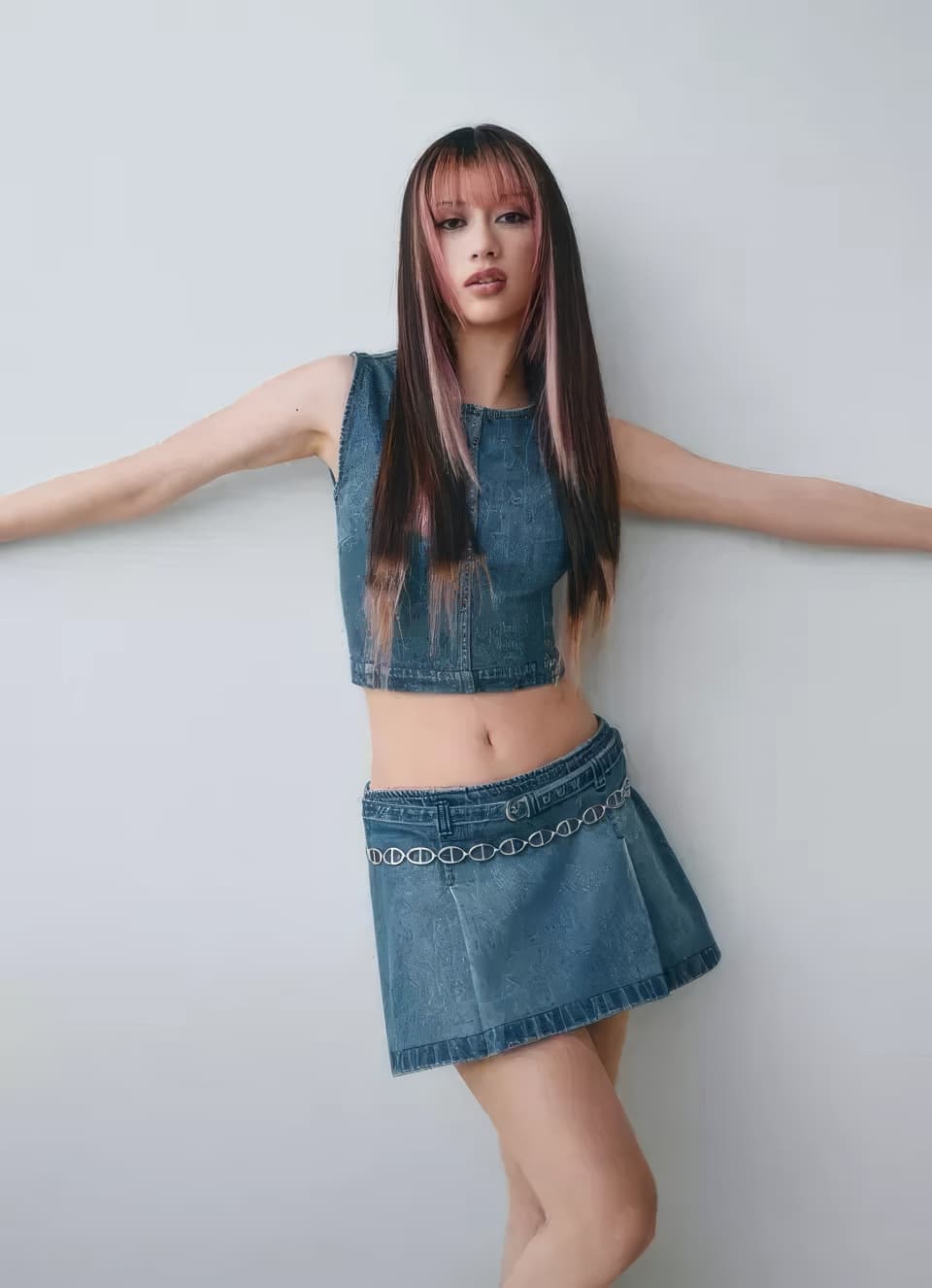 A young woman wearing a denim crop top and pleated denim skirt posing against a neutral grey wall.