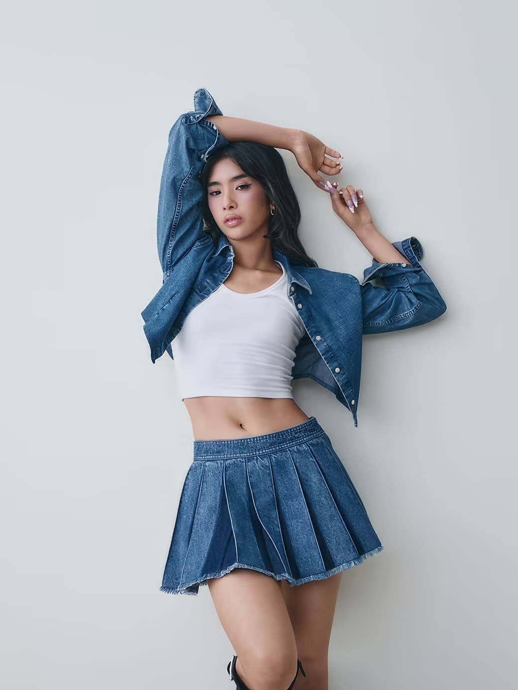 Young woman wearing a denim jacket, white crop top, and pleated denim skirt posing against a plain white wall