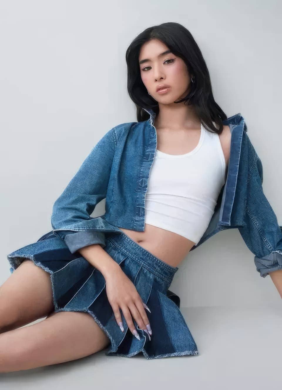 Model wearing denim jacket, white tank top and pleated denim skirt against a grey seamless studio background.