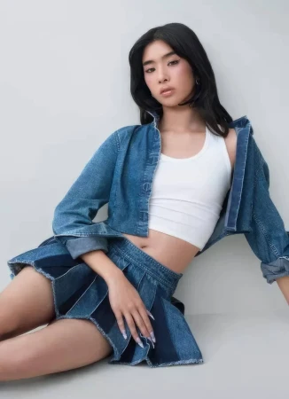 Model wearing denim jacket, white tank top and pleated denim skirt against a grey seamless studio background.