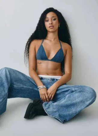 Woman with long dark curly hair wearing a blue denim halter top and jeans sitting against a white wall