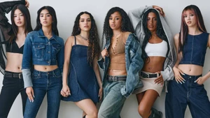Group of six young women posing in casual denim and streetwear against a neutral light grey studio backdrop.