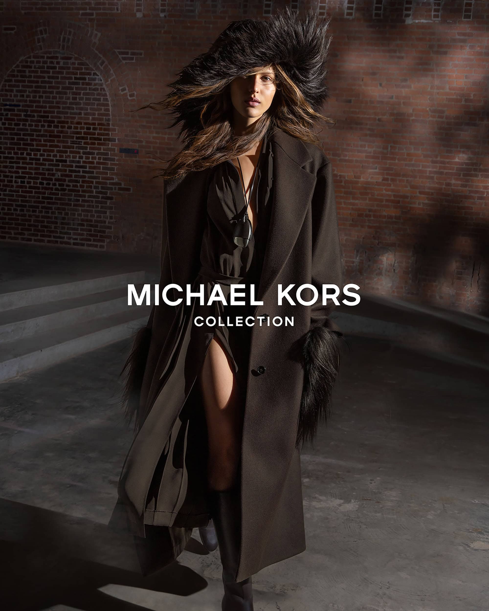 Model Angelina Kendall in a dark coat and faux fur hat standing before a brick wall for Michael Kors Collection Fall 2025.