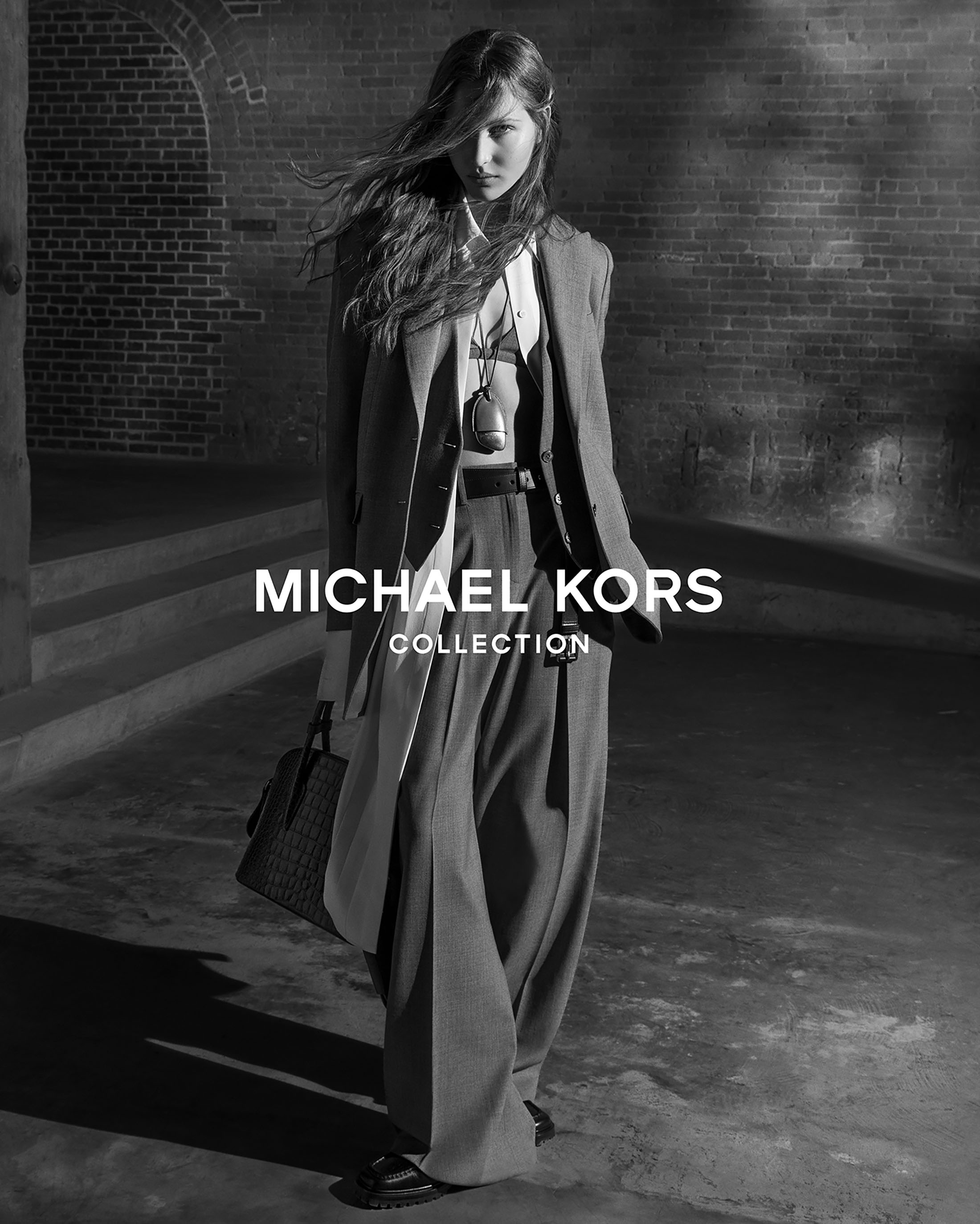 Model Angelina Kendall in tailored trousers and blazer holding a handbag against a brick wall for Michael Kors Collection Fall 2025.