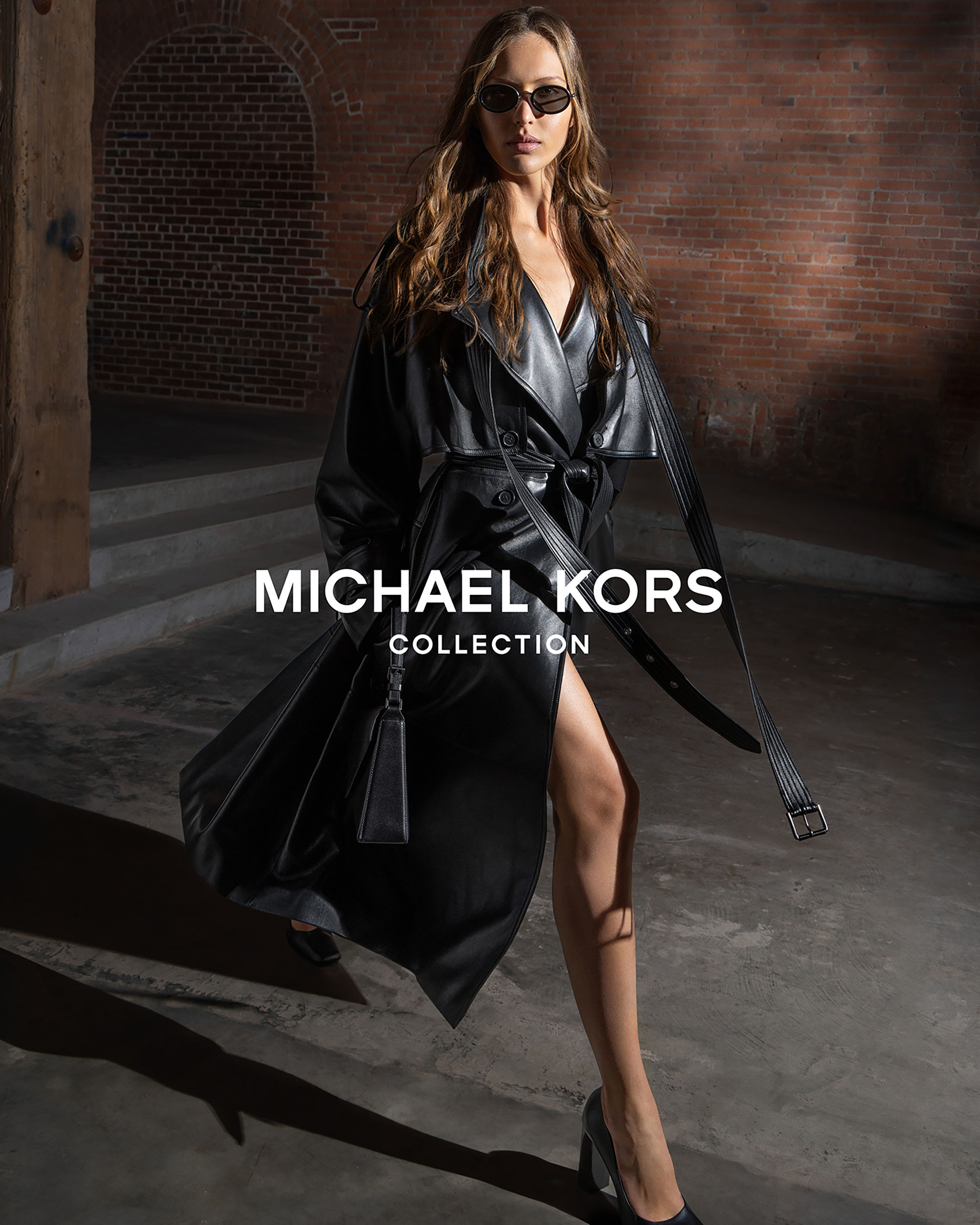 Model Angelina Kendall in a black leather trench coat walking inside Terminal Warehouse for Michael Kors Collection