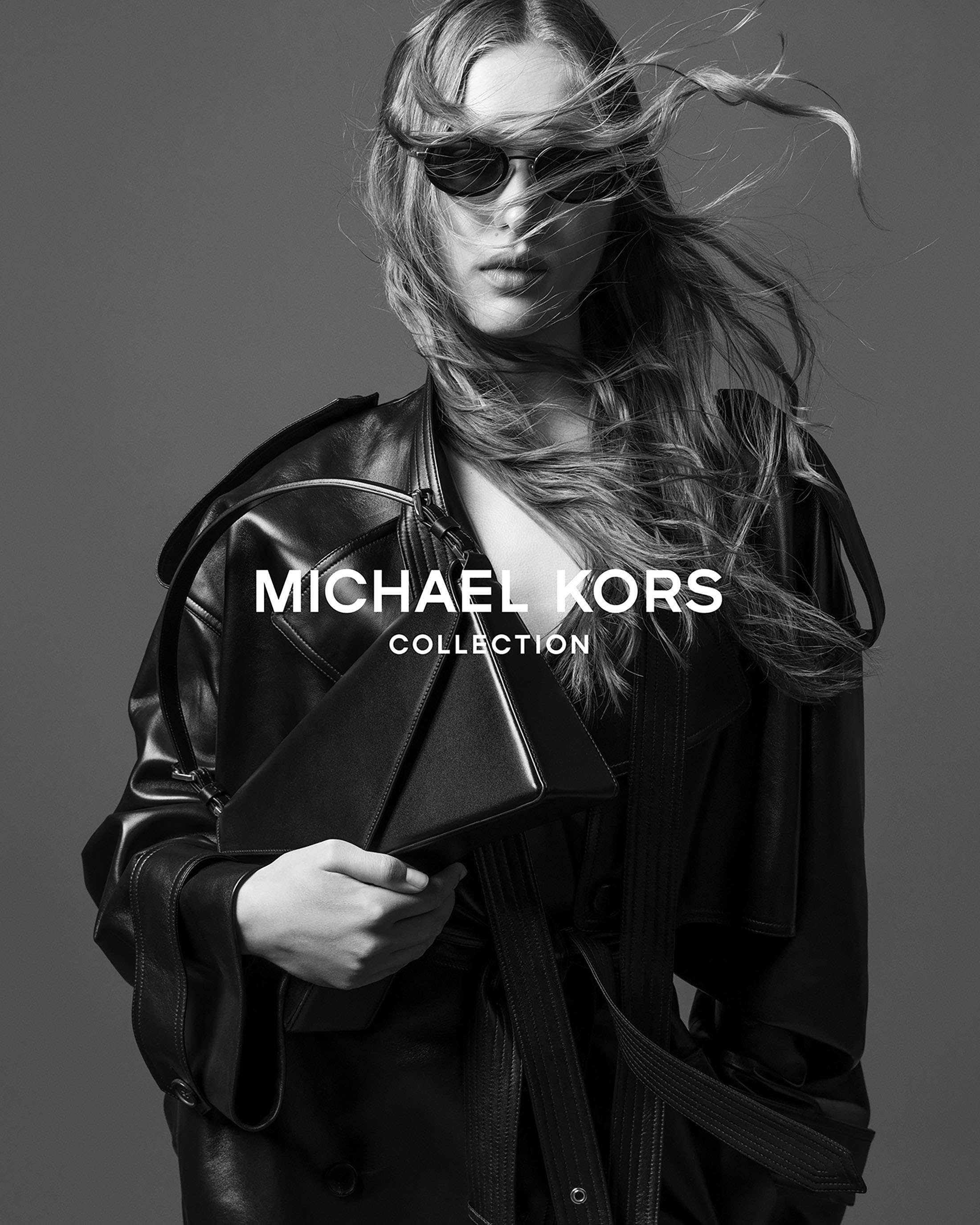 Model wearing dark sunglasses and a black leather coat holding a black handbag against a grey backdrop.