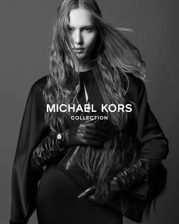 Model in black cape and leather gloves holding a dark handbag against a grey studio backdrop for Michael Kors Collection Fall 2025 ad campaign.