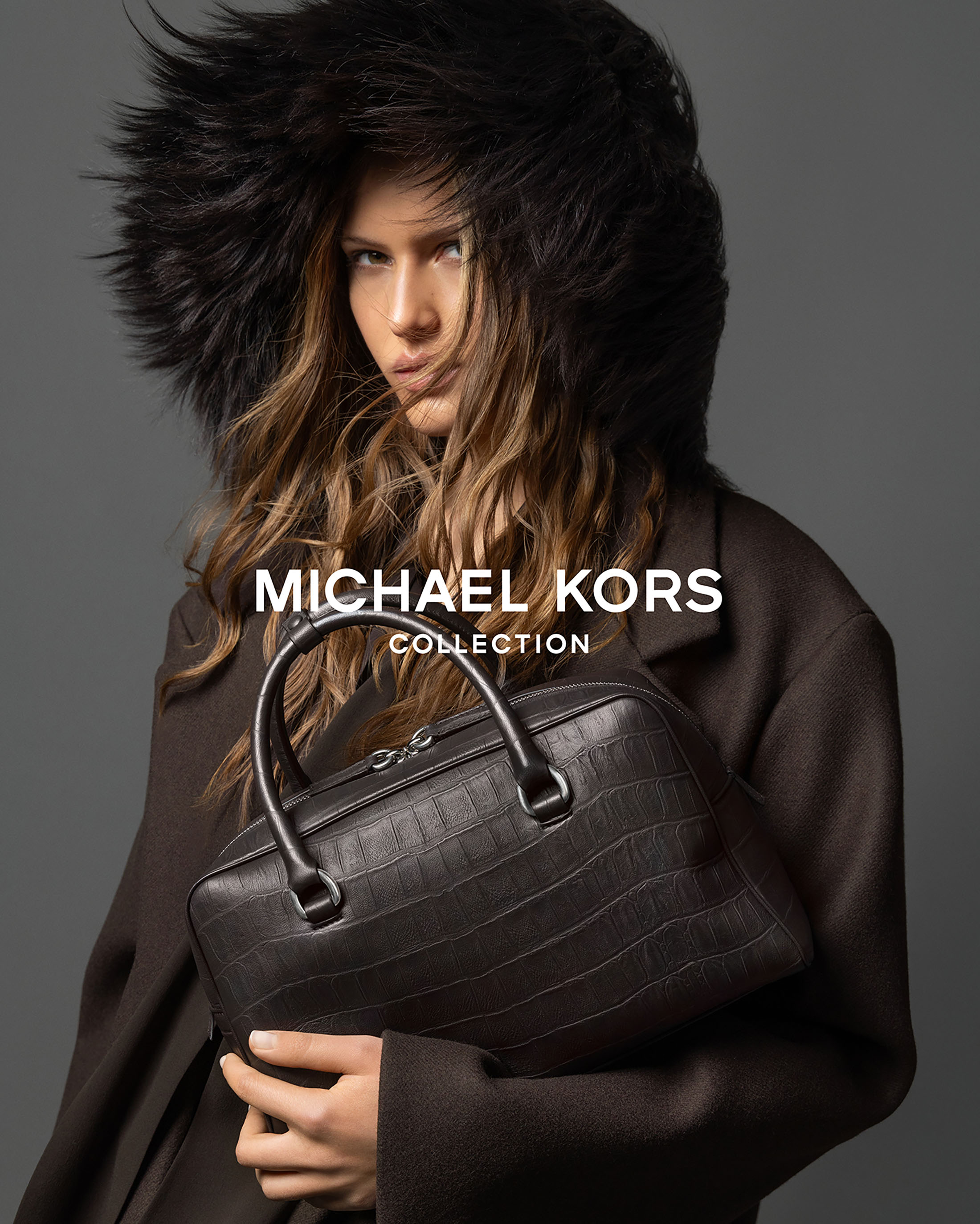 Model wearing a dark fur-trimmed hood and brown coat while holding a dark structured leather handbag against a solid grey background