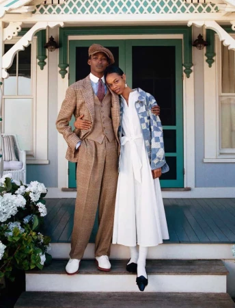 A man in a plaid tweed suit and newsboy cap posing with a woman in a white dress and patterned jacket on a porch.