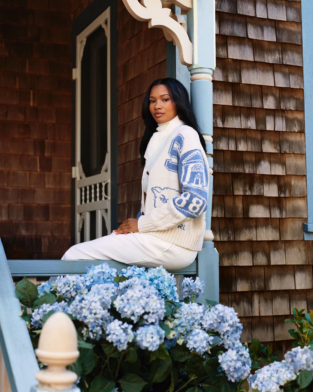 Model wearing a white and blue cardigan seated on a porch railing with blue hydrangeas in the foreground.