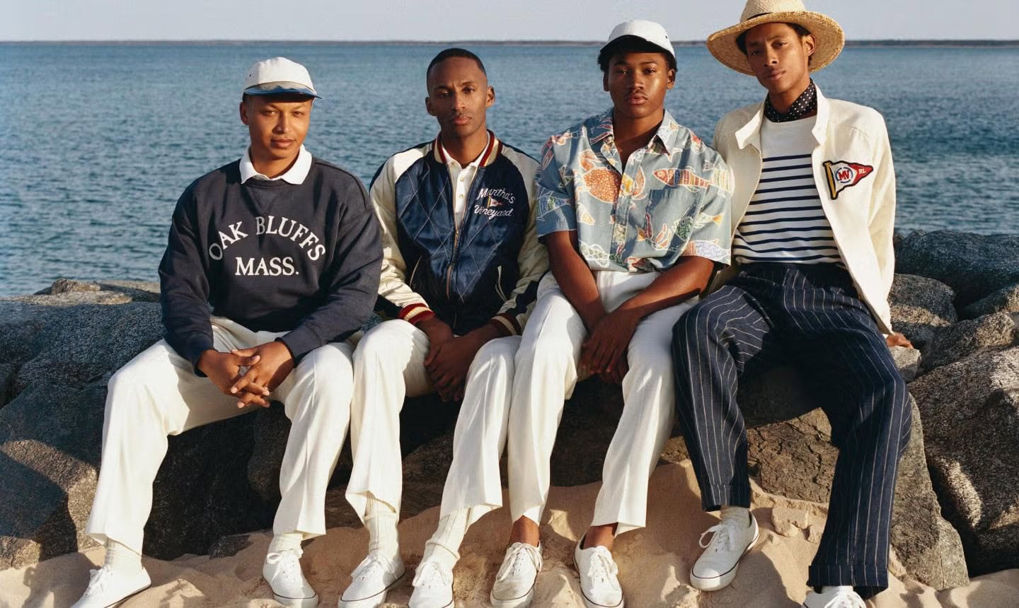 Four young men sitting on rocky shore wearing casual preppy apparel with water in the background