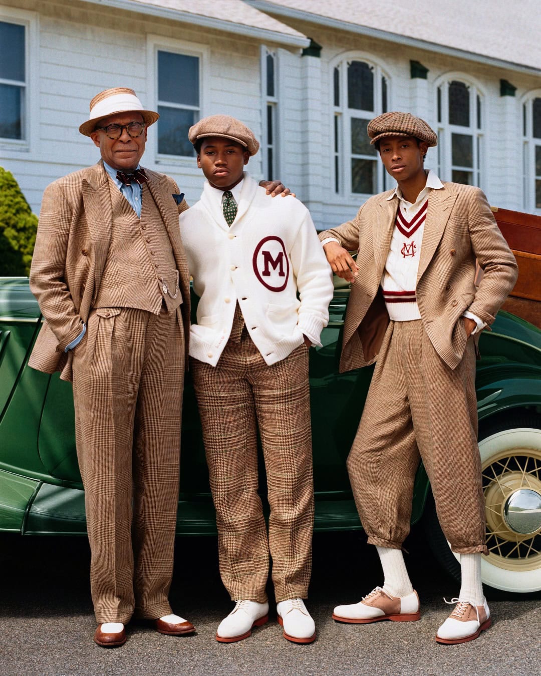 Three men in classic tailoring pose beside a vintage green car in front of a white building.