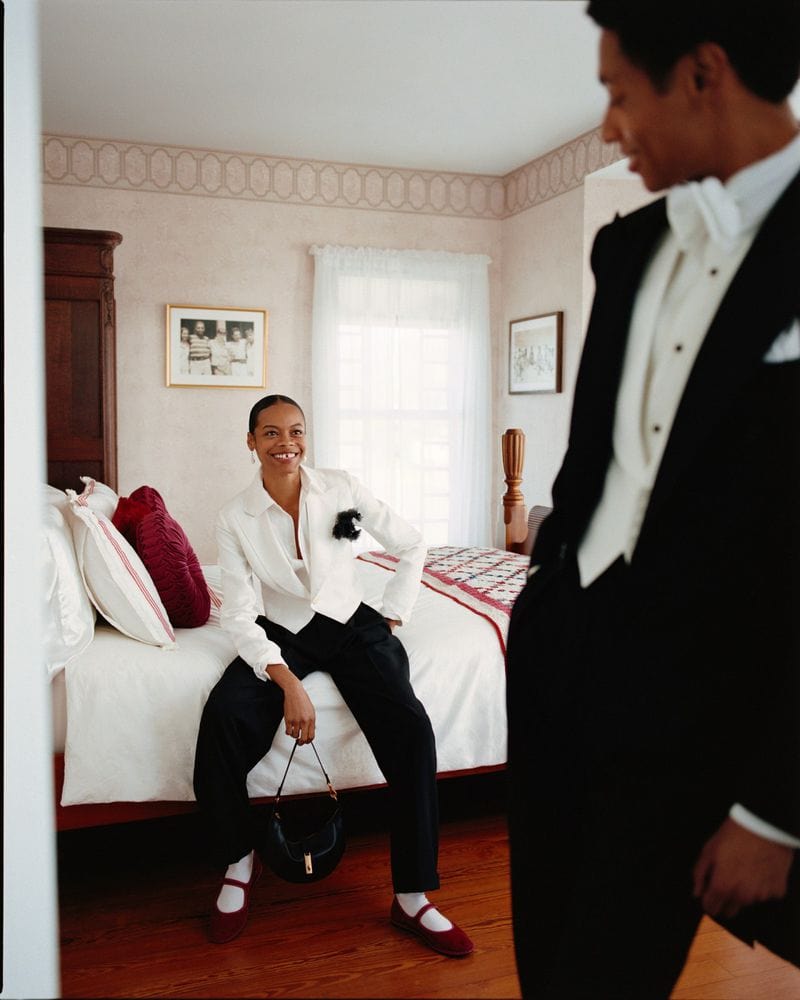 Model seated on a bed in a white shirt and black trousers, holding a handbag, while a man in a tuxedo stands in the foreground.