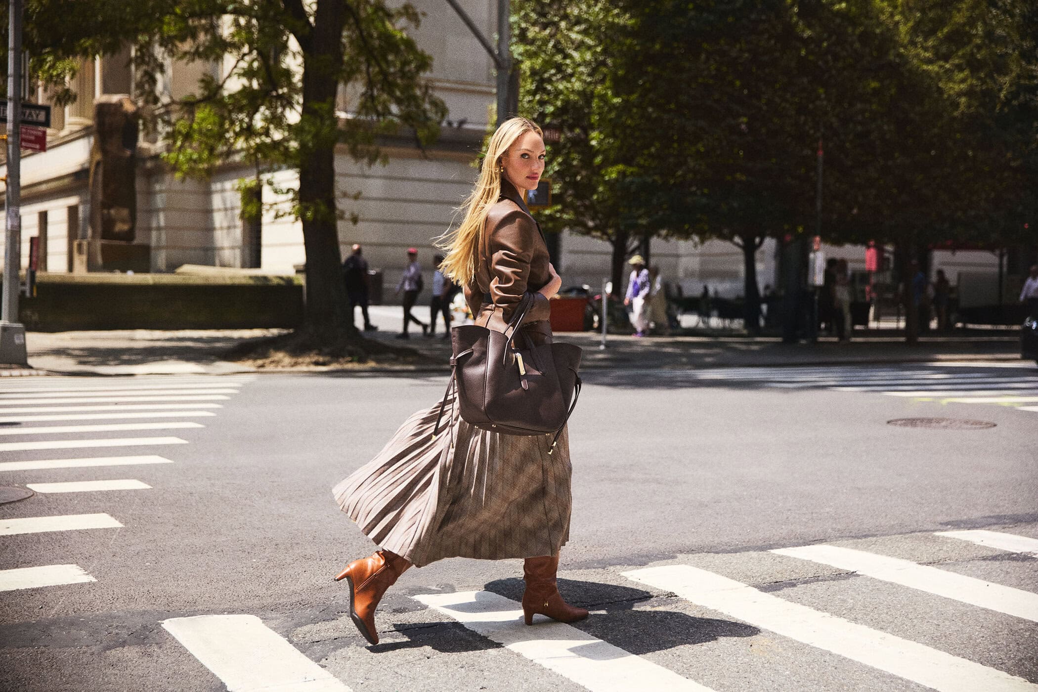 Candice Swanepoel crossing city street wearing brown leather jacket and pleated skirt carrying handbag