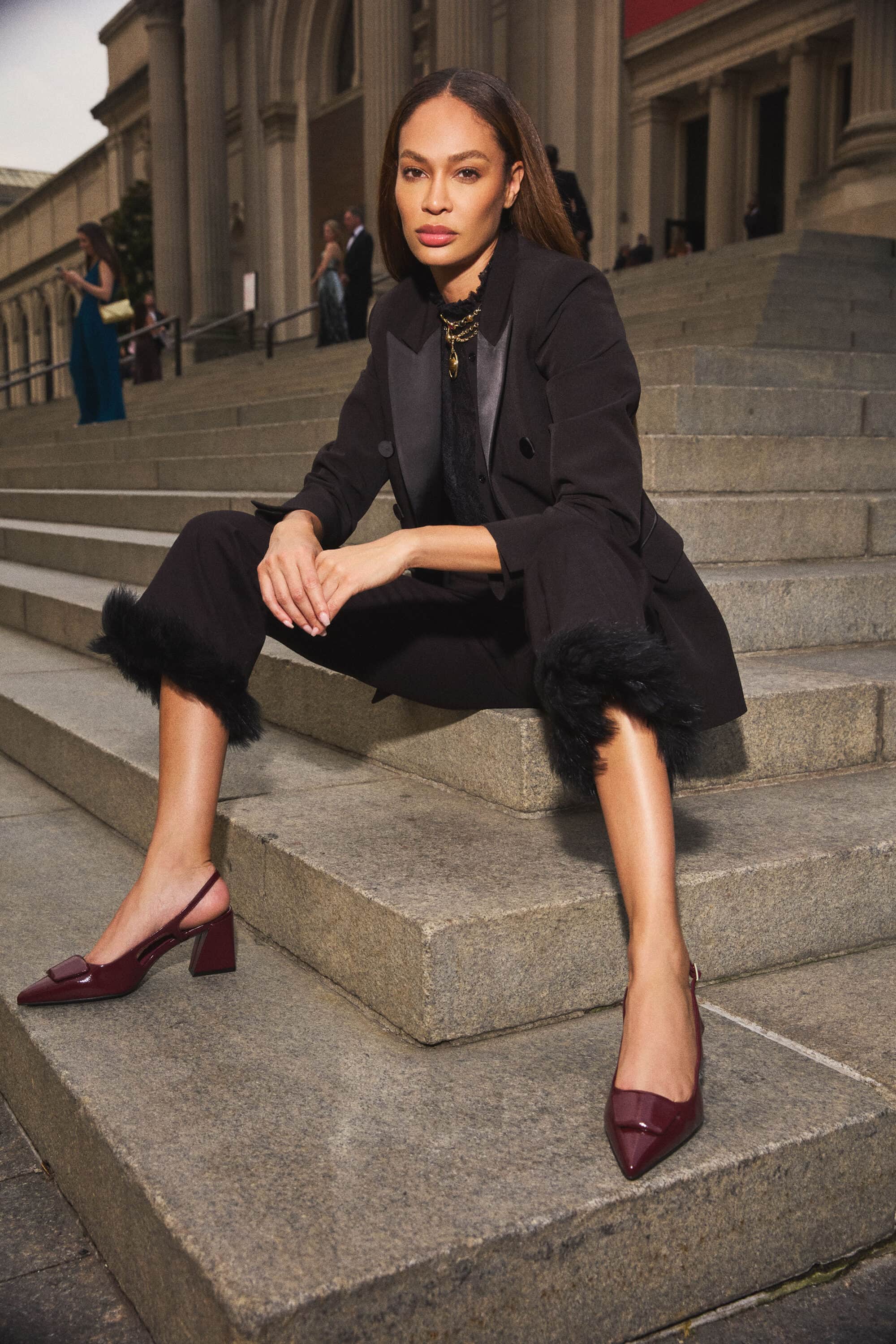 Joan Smalls seated on grand stone steps wearing a black blazer and trousers with dark heeled shoes for Anne Klein.