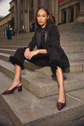 Joan Smalls seated on grand stone steps wearing a black blazer and trousers with dark heeled shoes for Anne Klein.