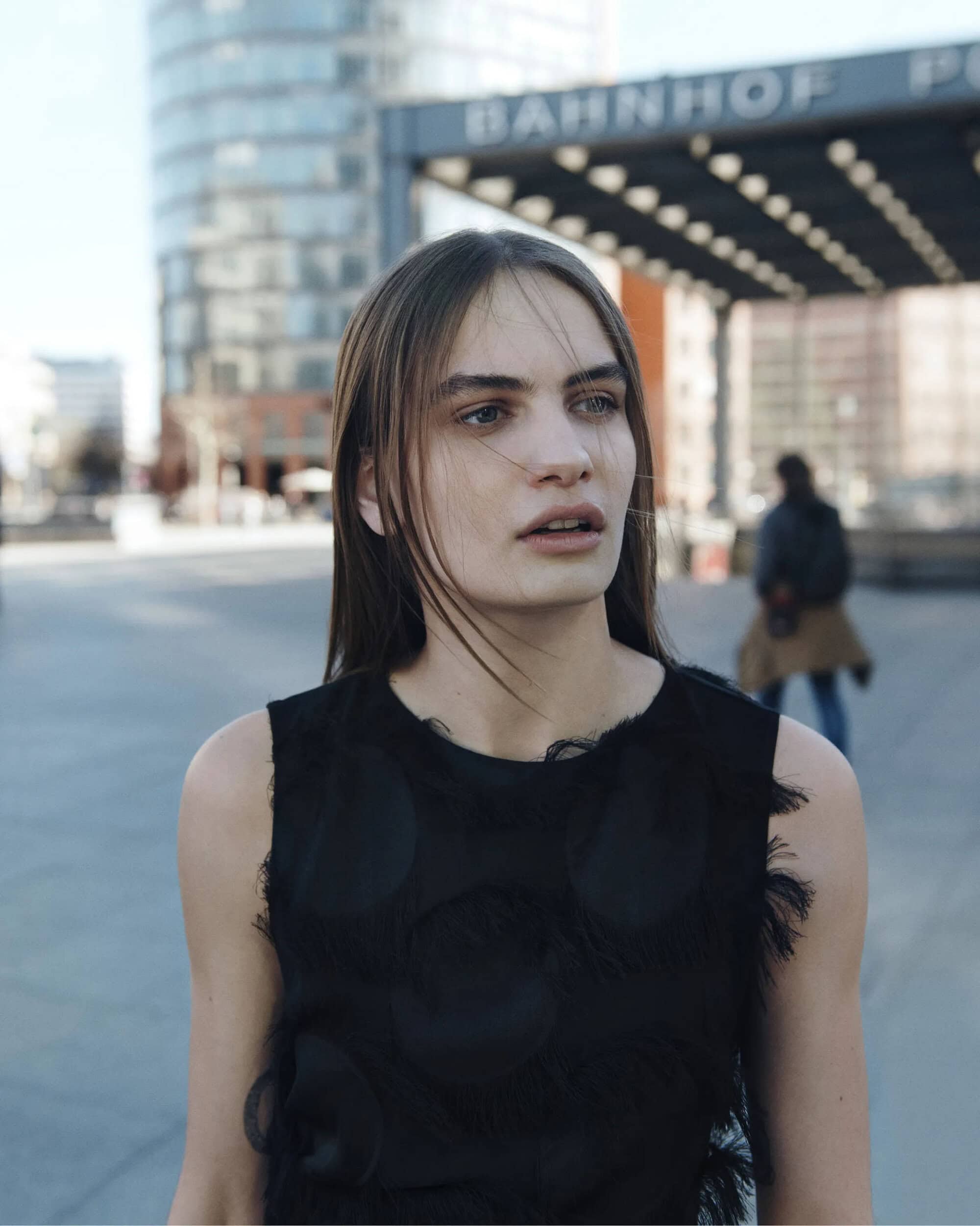 Close-up of a young woman wearing a black sleeveless top with fringed details outdoors against an urban background.