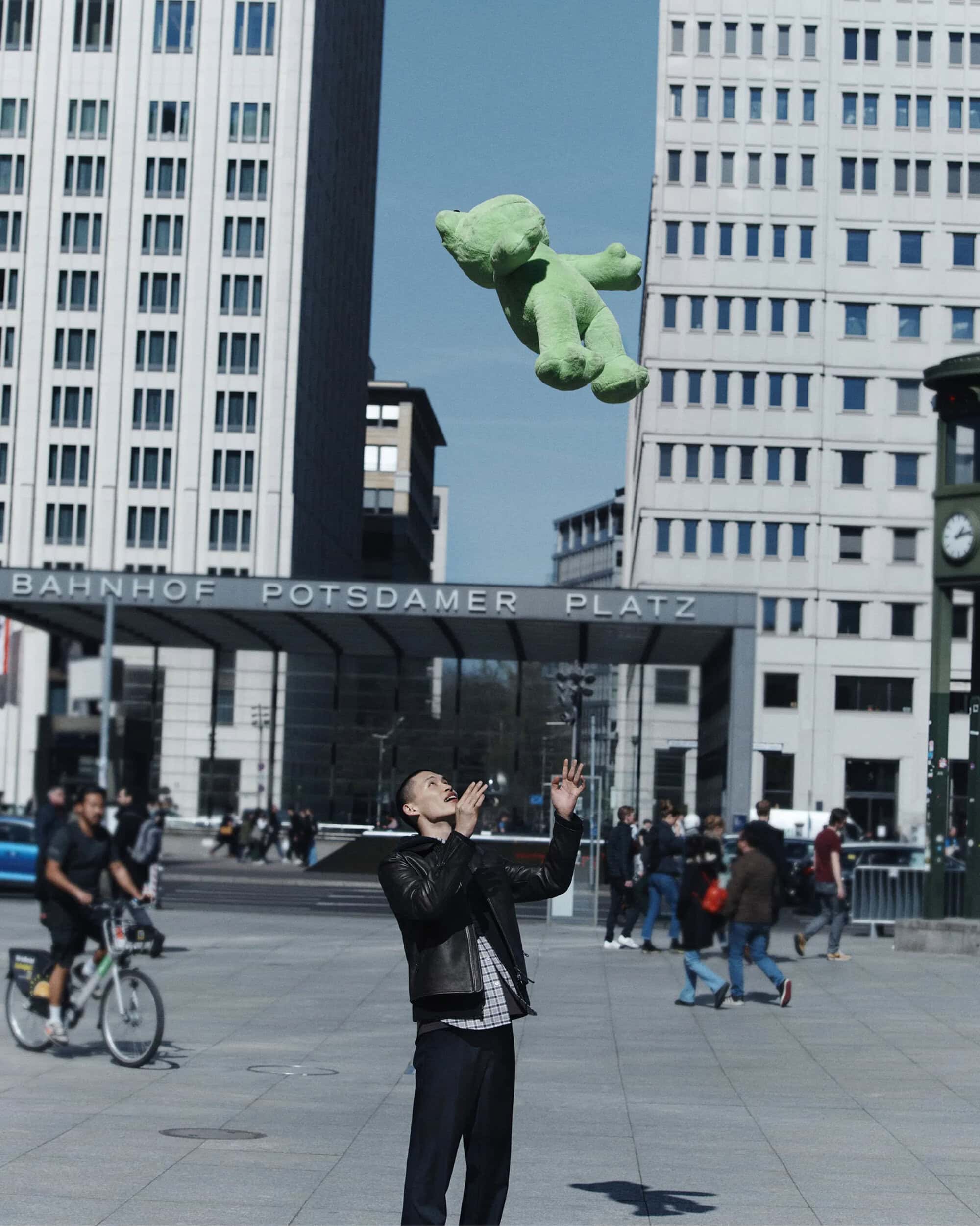 A man in a black jacket stands in an urban plaza tossing a large green stuffed bear into the air.