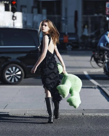 Young woman in a black fringe dress holding a bright green plush toy while walking across a city street