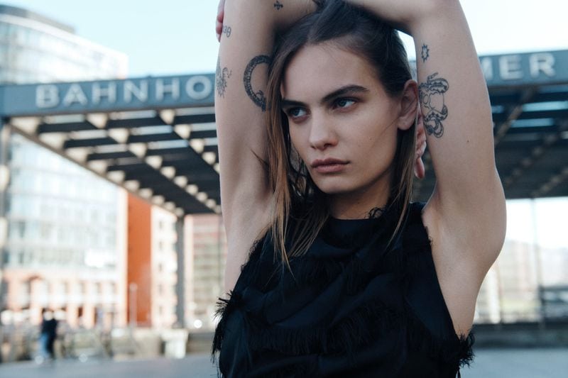 Model with tattooed arms wearing a black fringed top outdoors in front of a modern urban structure.