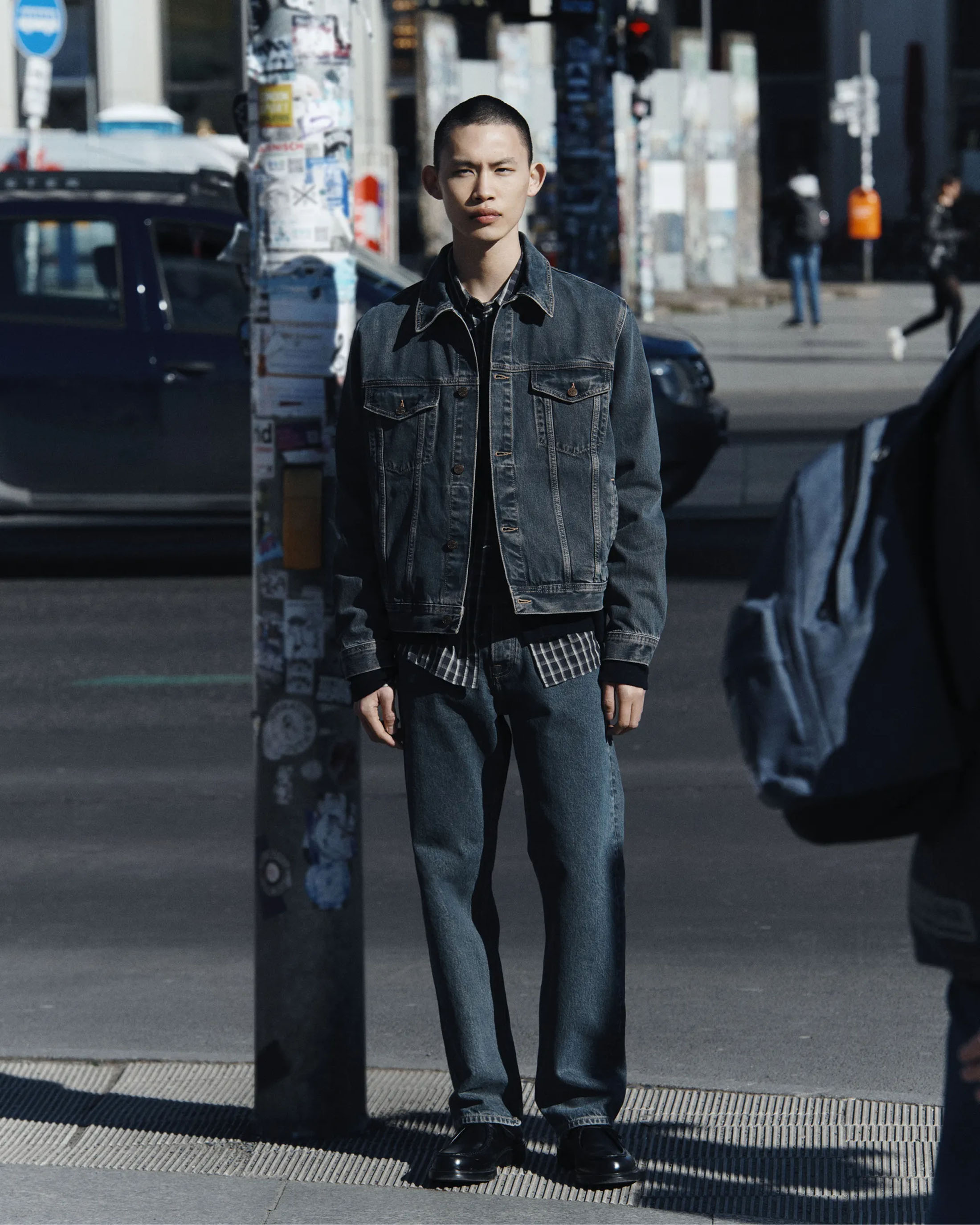 Young man standing on an urban street wearing a dark denim jacket and matching denim trousers.