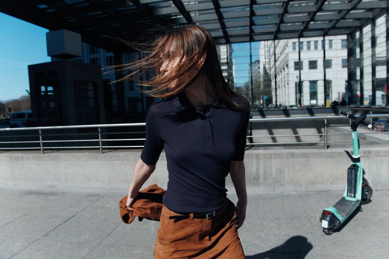 Woman in dark polo shirt and brown skirt with hair blowing in wind standing outdoors near scooter