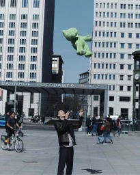 A man in a black jacket stands in an urban plaza tossing a large green stuffed bear into the air.