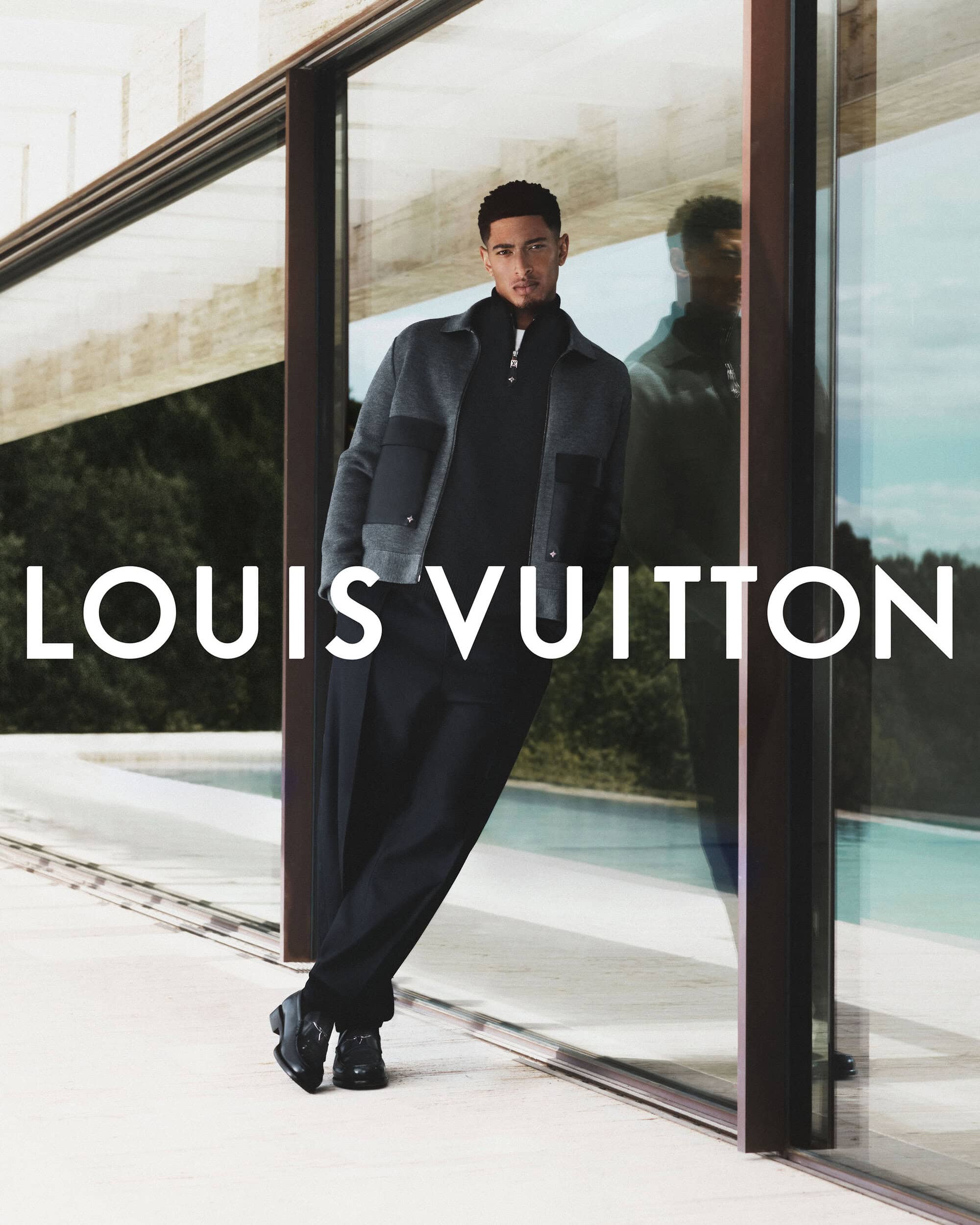 Jude Bellingham posing in a grey jacket and dark trousers by a glass pool wall for Louis Vuitton