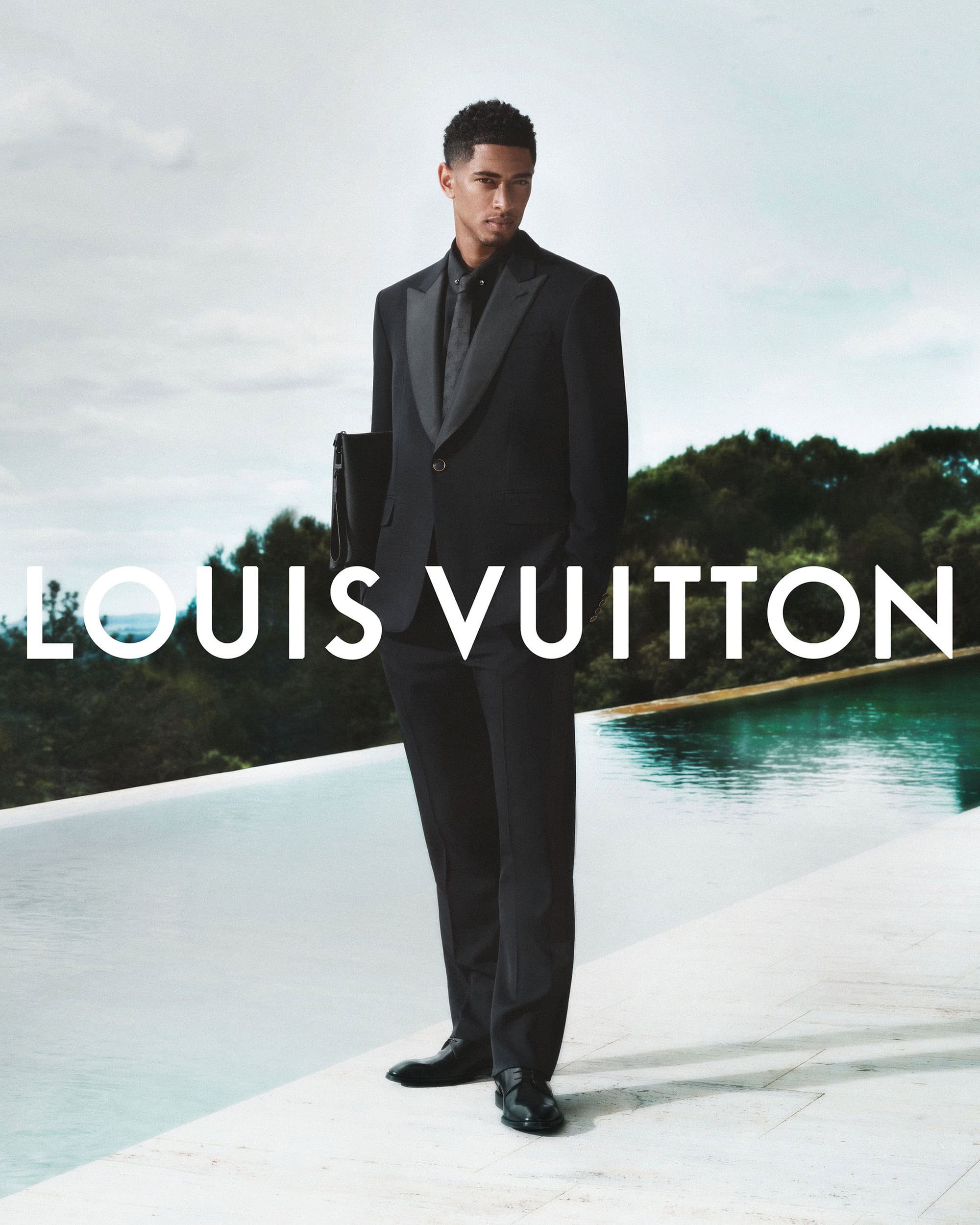 Man wearing a black suit standing by a pool outdoors with Louis Vuitton text overlay