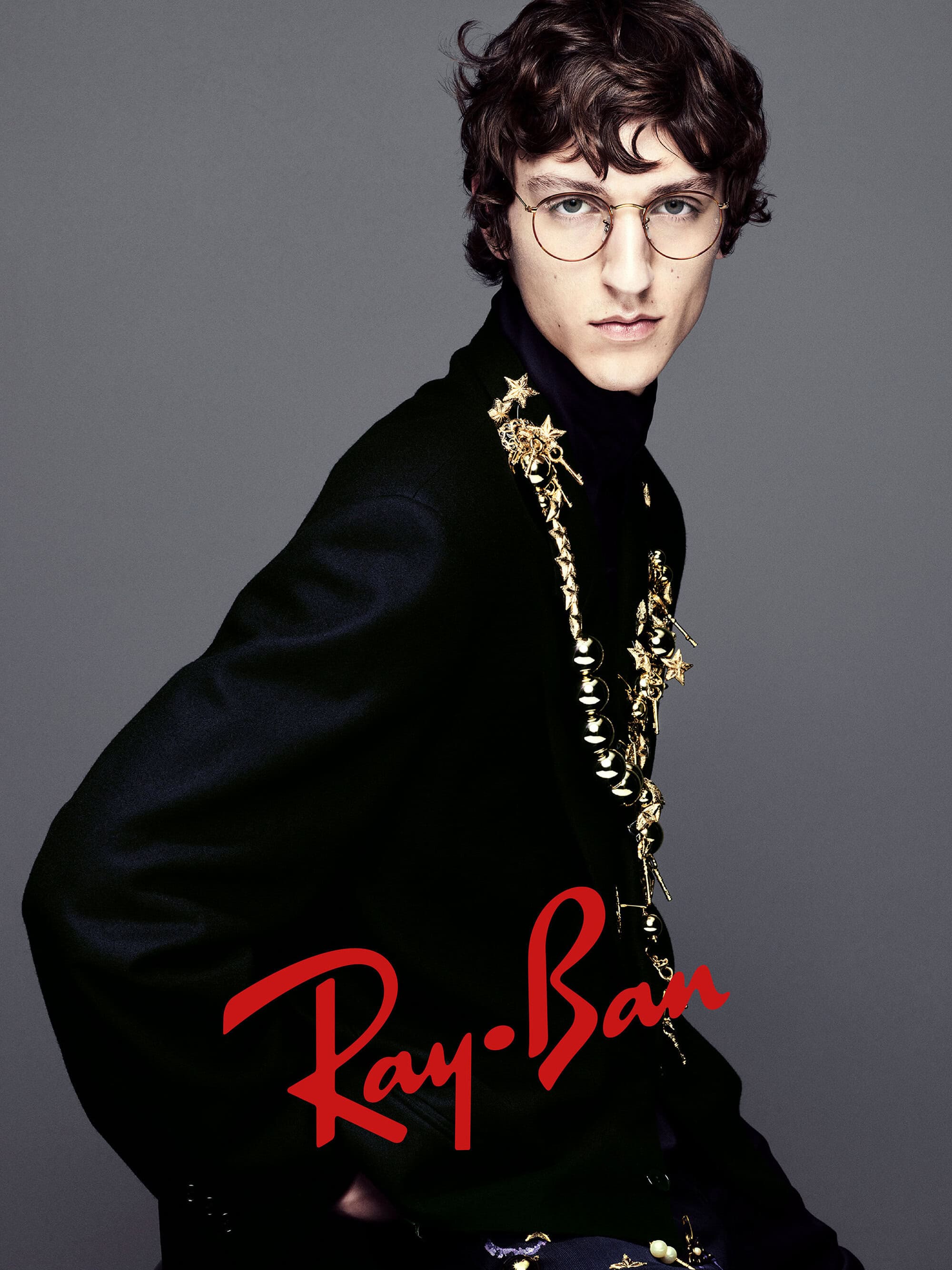 A man wearing round glasses and a black jacket adorned with gold charms against a grey studio backdrop.