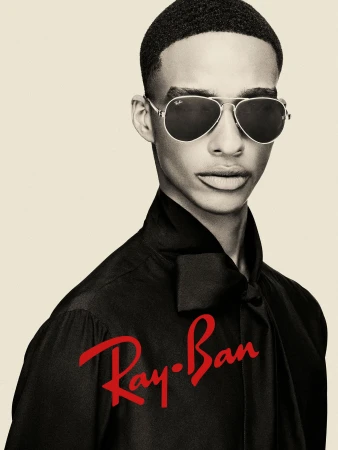Model wearing Ray-Ban aviator sunglasses and a black silk shirt against a neutral background with red Ray-Ban logo