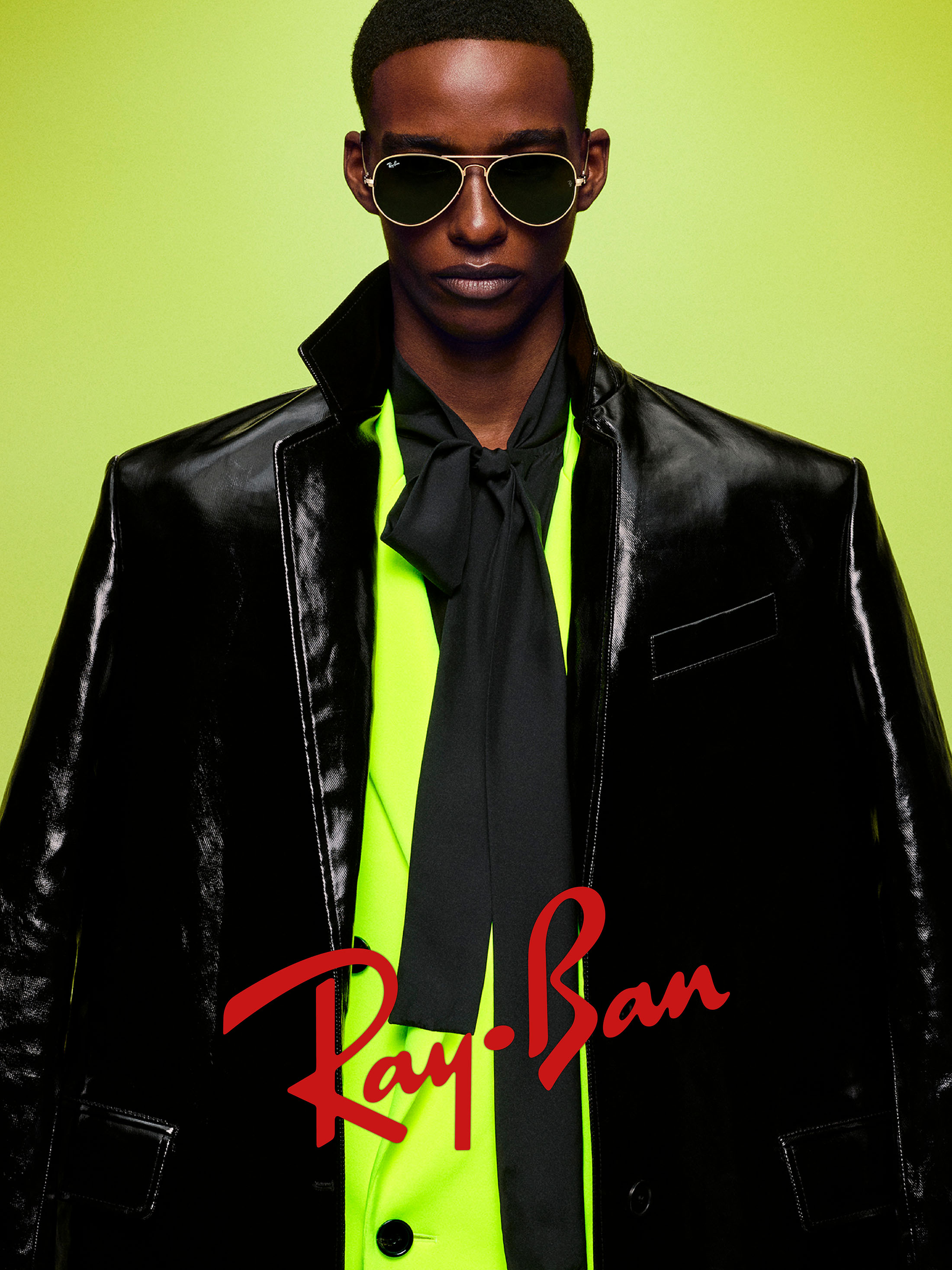 Man wearing aviator sunglasses, black coat, and lime green shirt with a tie against a bright backdrop for Ray-Ban The Icons 2025 campaign