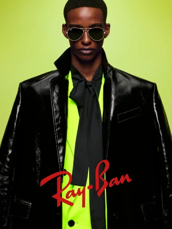 Man wearing aviator sunglasses, black coat, and lime green shirt with a tie against a bright backdrop for Ray-Ban The Icons 2025 campaign
