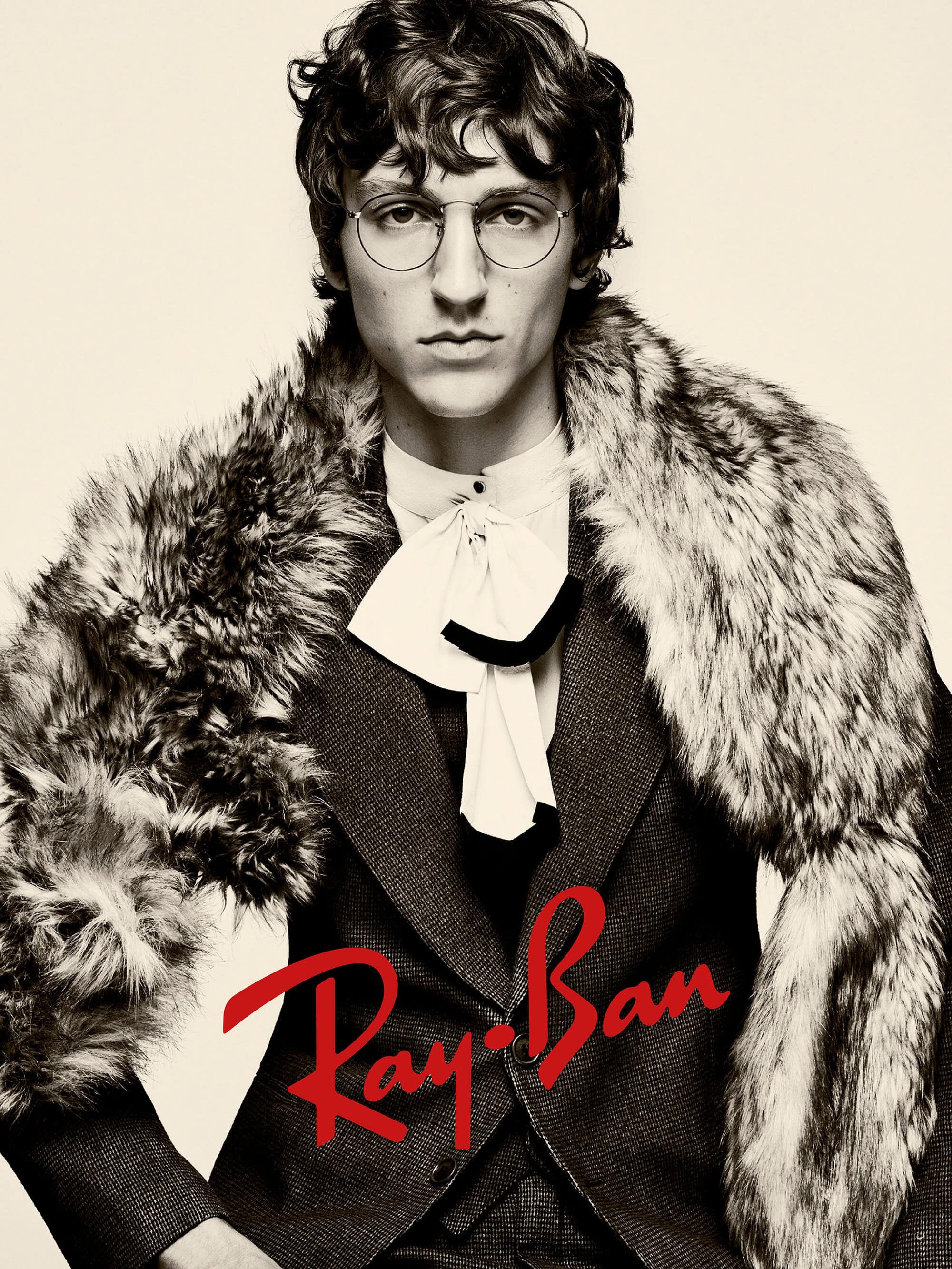 Close-up portrait of a man wearing round glasses, a dark blazer, and a fur stole against a pale background