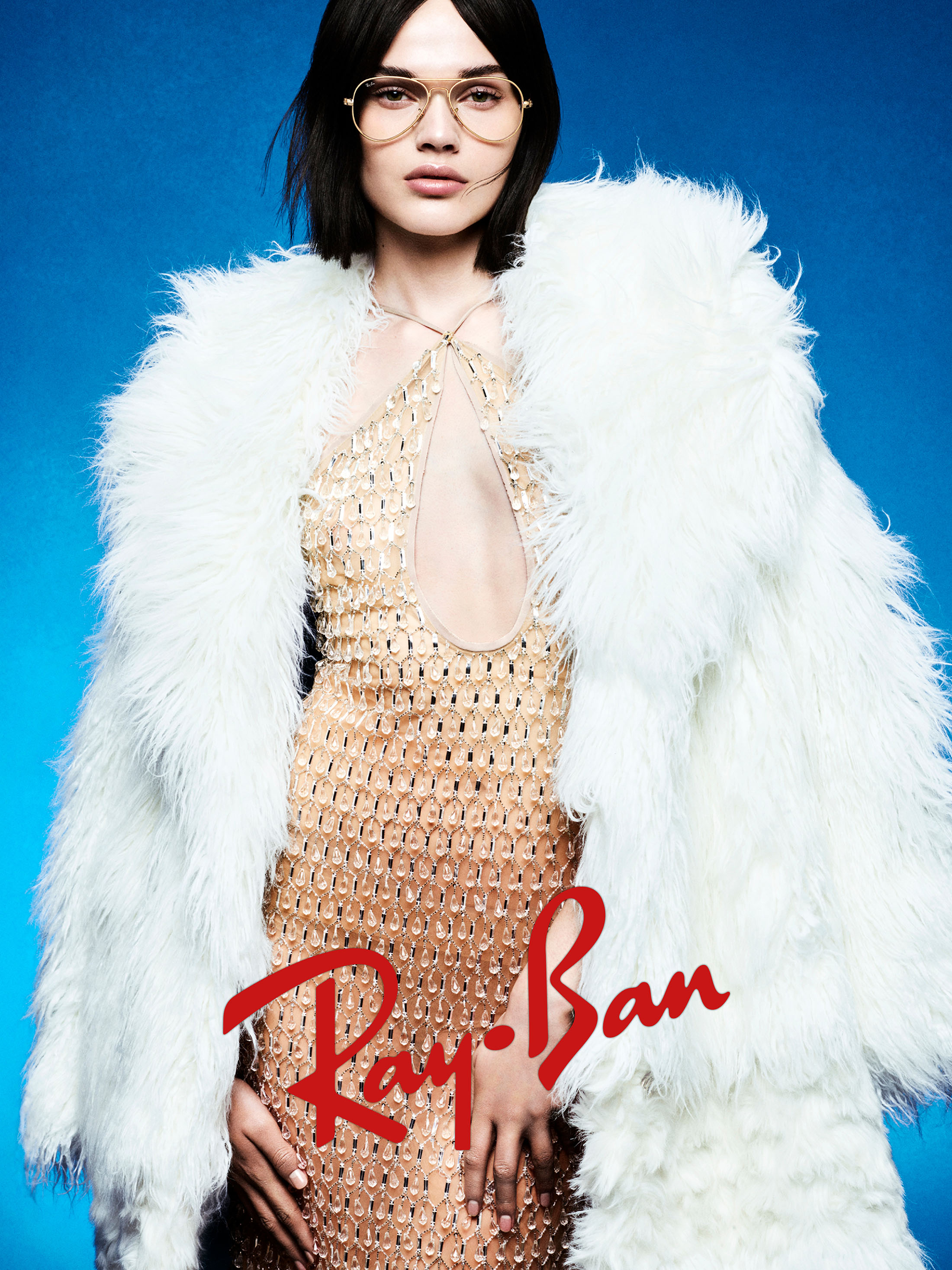 Model wearing gold wireframe glasses and a fluffy white coat over a metallic dress against a bright blue background