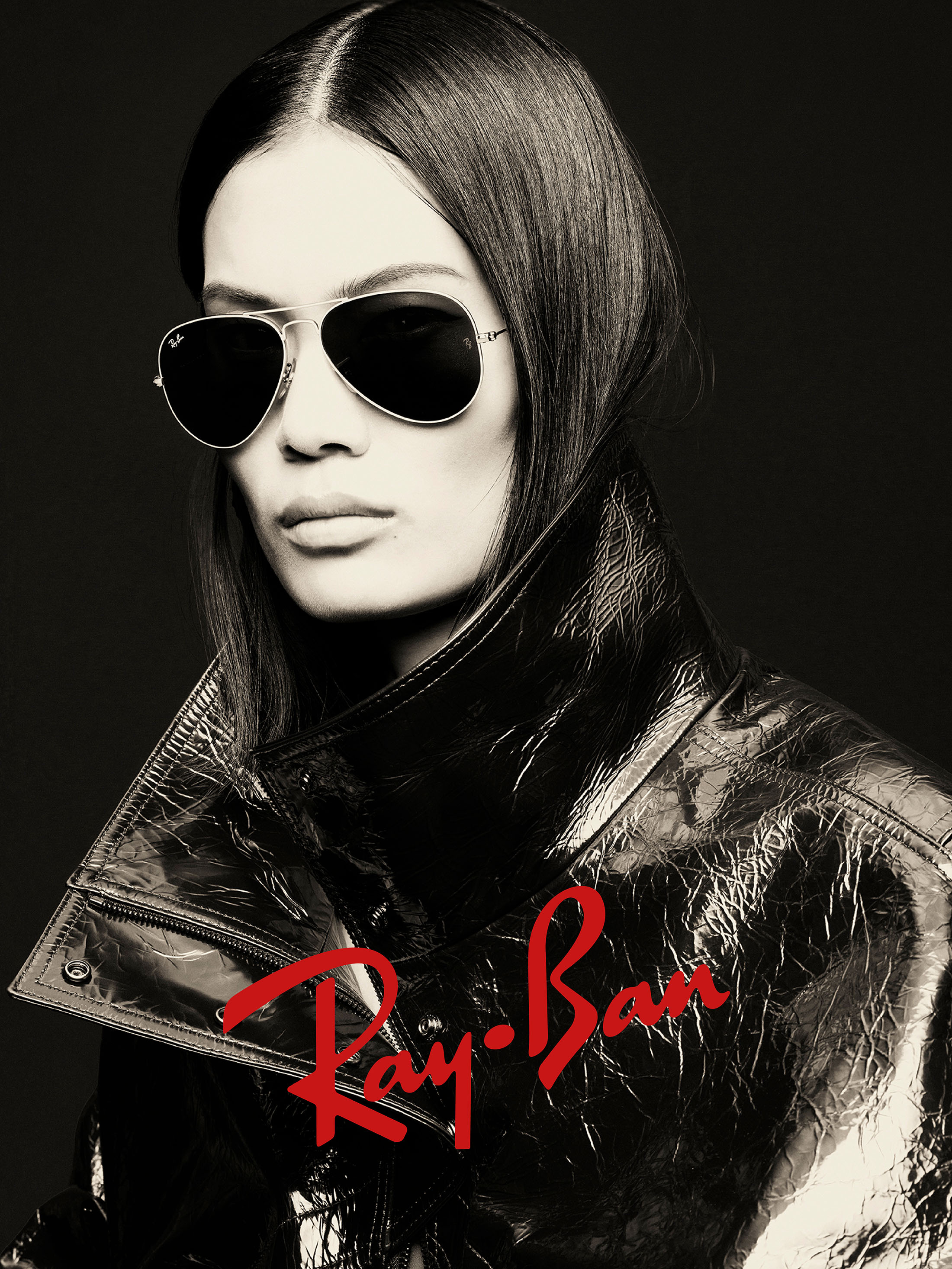 Model wearing dark aviator sunglasses and a shiny metallic jacket against a black background with a red Ray-Ban logo